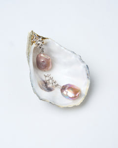 A pair of square baroque pearl earrings in pink, lavender, and gold hues, set in silver-tone latch-back clasps, displayed in an oyster shell. Mayfair Pearls. 