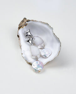 A pair of silver-colored earrings with a star-shaped clasp and a druzy stone, accompanied by a white coin pearl, displayed on an oyster shell. Mayfair Pearls jewellery