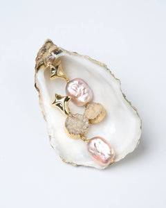 A pair of gold-tone starburst earrings with a natural druzy stone and pink baroque pearls displayed on a white oyster shell.