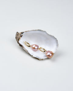 A pair of blush pink baroque pearl earrings with gold twist clasp, displayed on a white oyster shell.