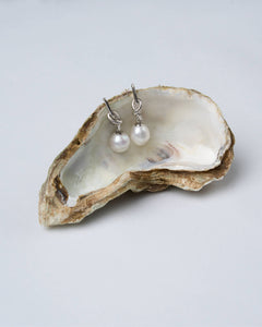 A pair of twist knot petite pearl earrings, with silver-tone latch-back clasps, displayed on a textured surface.