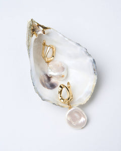 A pair of gold baroque pearl earrings with latch-back clasps displayed on a white oyster shell.
