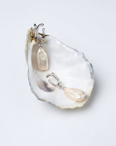 A pair of white pearl earrings with a curved design, displayed on a silver-tone latch-back clasp, presented on a white oyster shell.